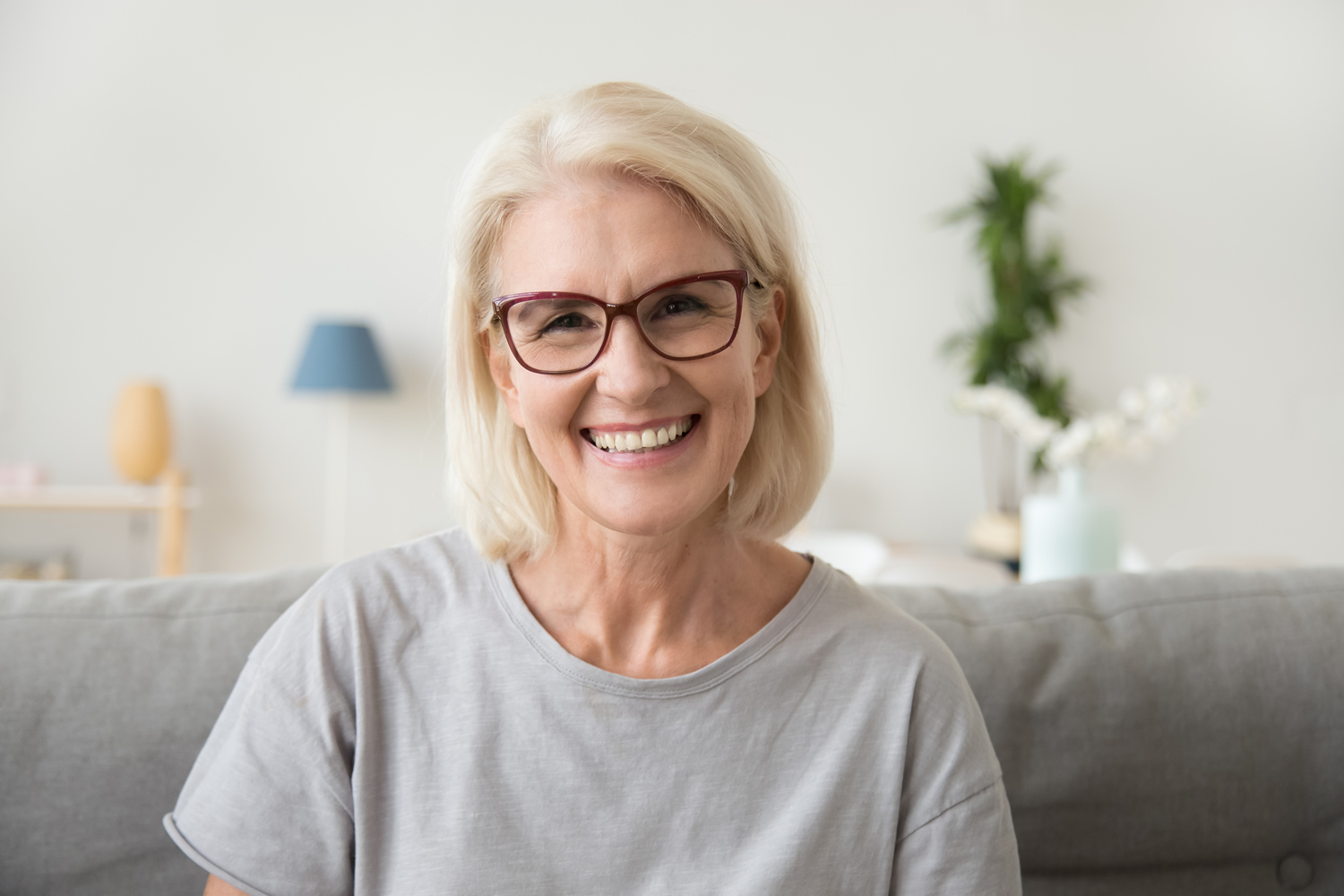 A smiling older woman with short blonde hair and glasses sitting on a couch in a bright living room.
