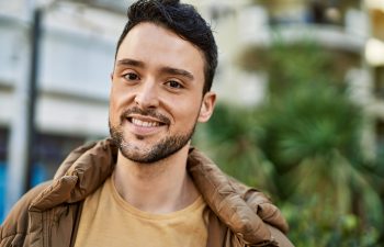 A young man with dark hair and a beard, wearing a brown jacket and beige shirt, stands outdoors and smiles at the camera with greenery and buildings in the background.