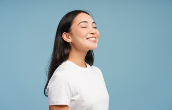 A young woman with long, dark hair, wearing a white t-shirt, smiles contentedly with her eyes closed, shown from the side against a plain blue background.
