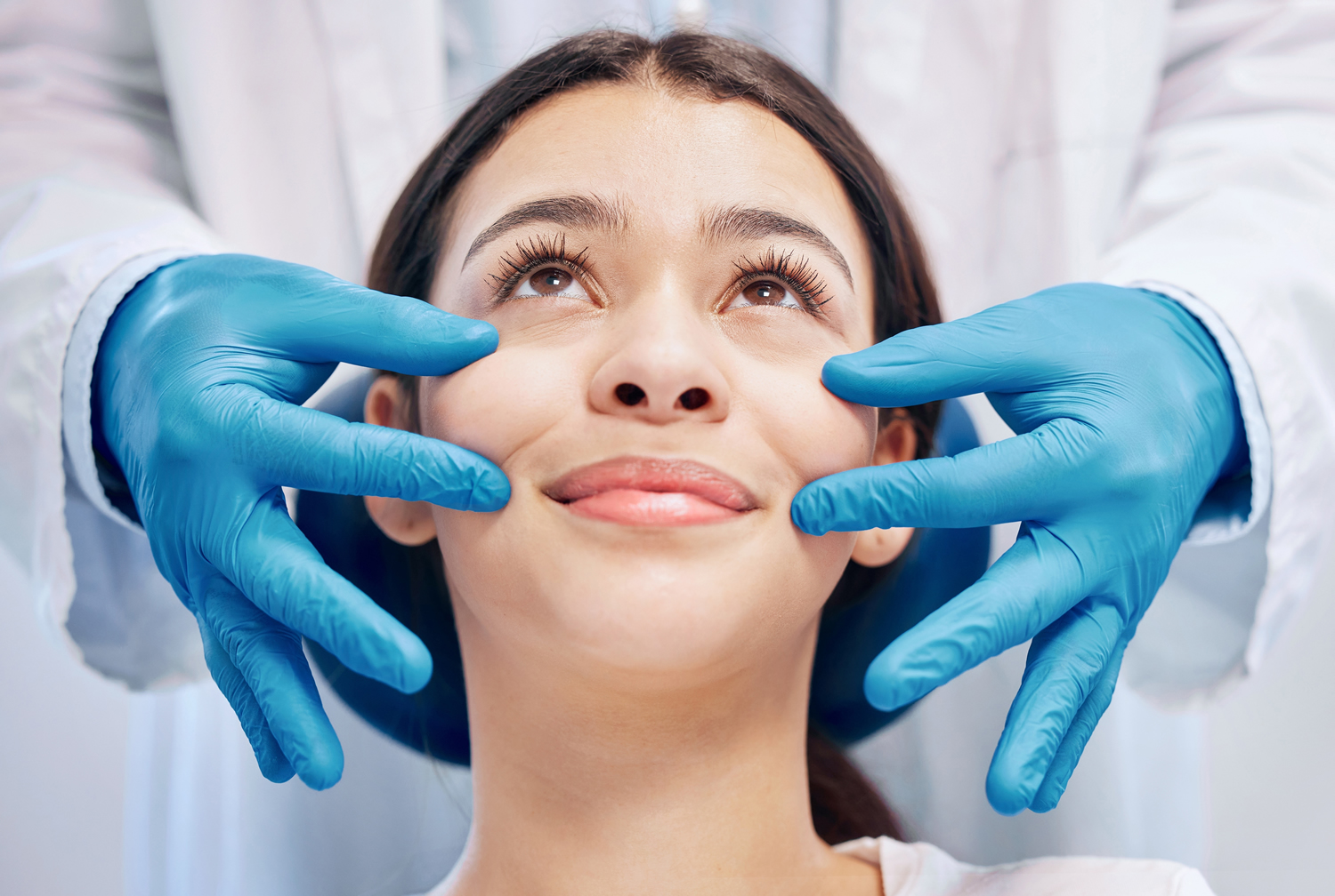 A medical professional in blue gloves gently touching the face of a smiling woman during a skin consultation or aesthetic procedure.